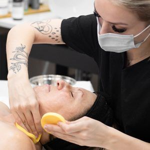 Blonde woman with white face mask and lash extensions applying yellow facial sponges to neck and cheek of female client laying down with closed eyes.