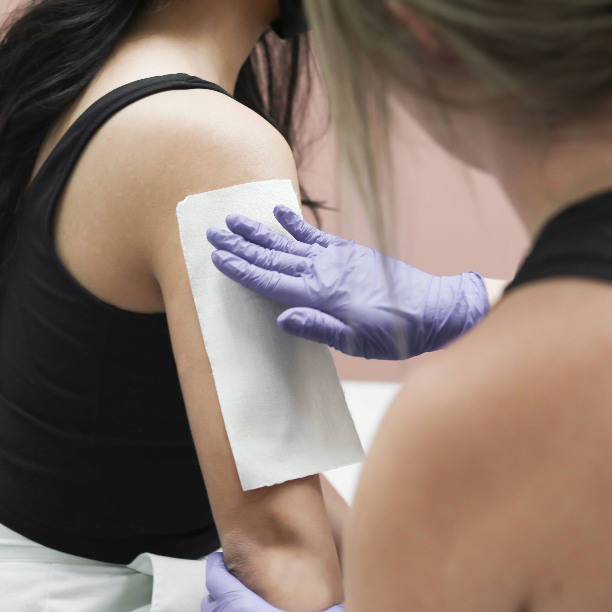 Student with gloved hand holding white wax strip against client's upper arm.