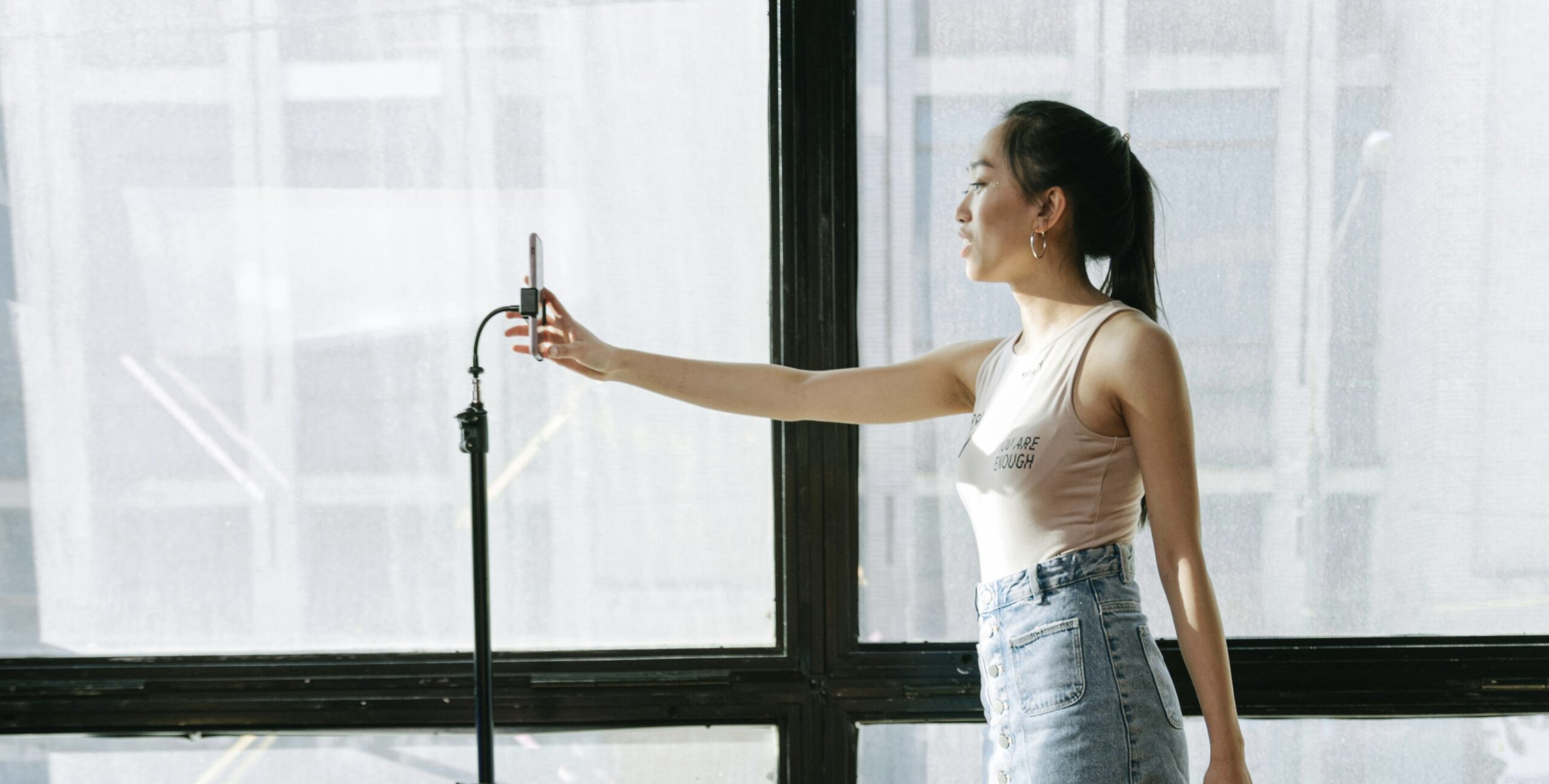 A young woman stands in front of large windows holding a smartphone mounted on a tripod, appearing to film herself. She is wearing a sleeveless top and high-waisted denim skirt, and is lit by natural daylight coming through sheer curtains. She is Optimizing Every Touchpoint 
