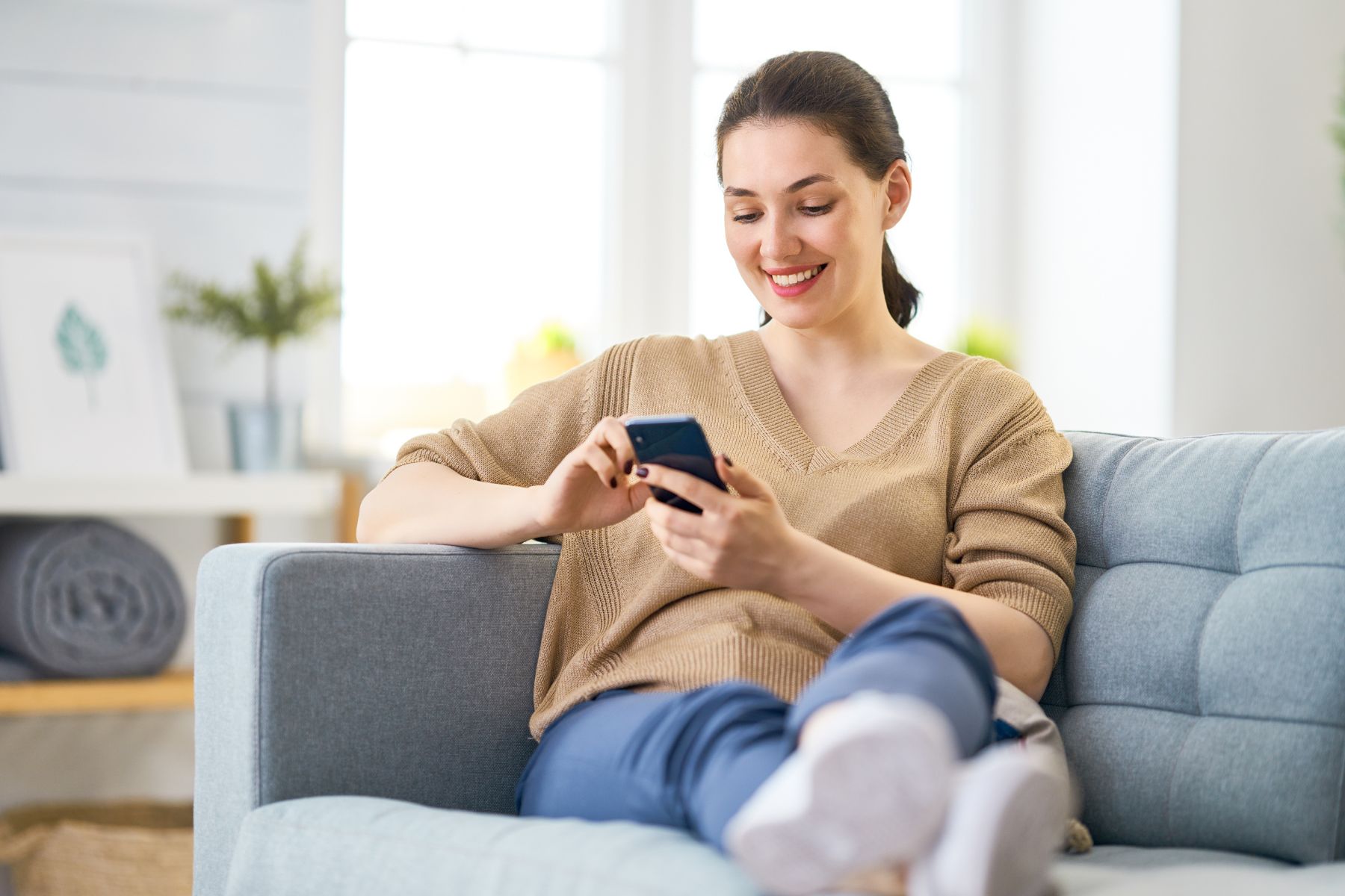 Smiling woman relaxing on a couch while using her smartphone, representing easy content creation in social media for estheticians