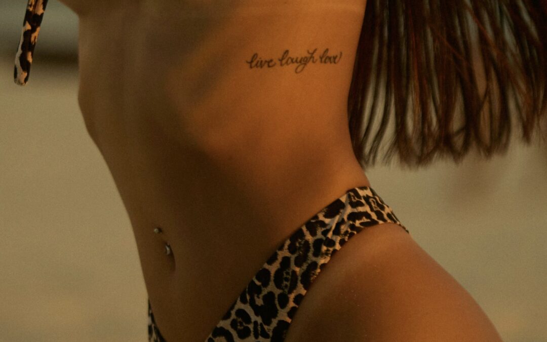 Side view of a woman in a leopard bikini with a script tattoo reading “Live Laugh Love,” symbolizing an outdated saying, tattoo regret, and the value of training through a laser tattoo removal course Winnipeg.
